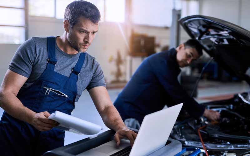 Mecânico usa checklist e fonte de informação técnica no notebook para orientar seu colega que está efetuando testes em um veículo parado.