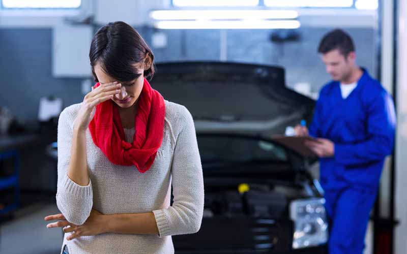 Cliente insatisfeita leva a mão à testa enquanto mecânico ao fundo refaz testes em um veículo parado